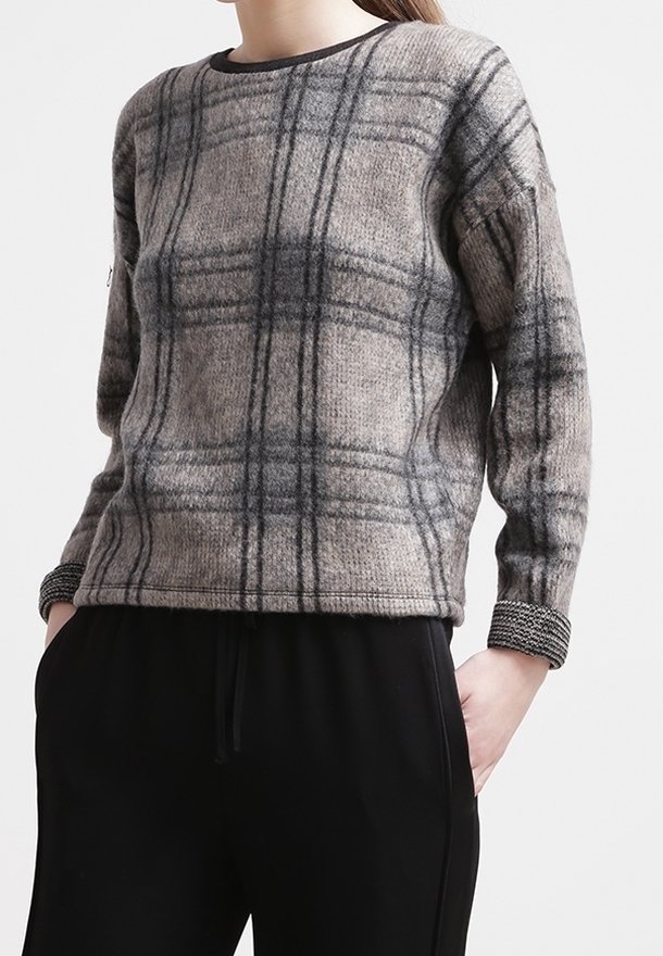 Femme portant un pull à manches longues à carreaux gris avec des poignets noirs et un pantalon noir, debout les mains dans les poches devant un fond uni.