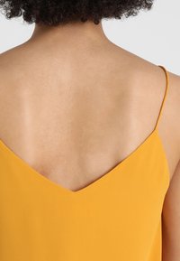 Žlutý tílko bez rukávů s výstřihem do V na zádech, vyrobené z hladké látky, s jednoduchým designem a tenkými ramínky.