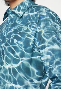 Camicia realizzata in tessuto con motivo d'acqua nelle tonalità di blu e acquamarina. Il design imita i riflessi della luce sull'acqua. Colletto con bottoni.