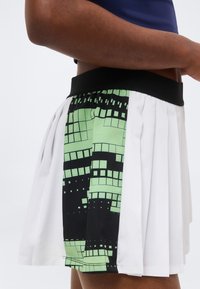 Κοντινό πλάνο ενός ατόμου που φοράει λευκή πλισέ φούστα με μαύρη και πράσινη σχεδιασμένη λωρίδα και μαύρη μέση.