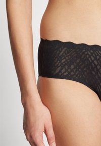 Culotte en dentelle noire avec un bord festonné, dotée d'un design texturé caractéristique de la dentelle fine. Le tissu semble doux et transparent.