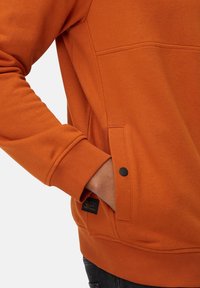 Orange sweatshirt lavet af struktureret stof med en sidelomme med en sort knap, ribbede manchetter og et mærke nederst til venstre.