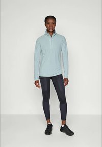 Giovane donna in piedi che indossa una felpa azzurro chiaro con zip, leggings neri con tasche e sneakers nere, contro uno sfondo chiaro e semplice.