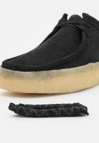 Black suede shoe with beige crepe sole, black laces, and separate bundled black shoelace on white background.