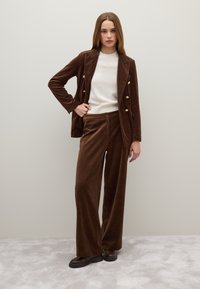 Completo in velluto a coste marrone composto da un blazer doppiopetto e pantaloni a gamba larga, abbinato a un maglione crema e calzature scure.