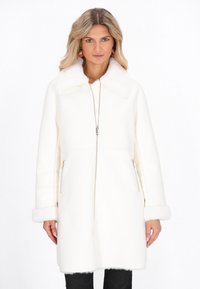 Vrouw in een witte middellange jas met bontkraag en -manchetten, voorrits en ritssluitingen aan de zijkanten, staande voor een witte achtergrond.
