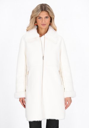 Donna che indossa un cappotto bianco di mezza lunghezza con colletto e polsini di pelliccia, cerniera frontale e tasche laterali con zip, in piedi davanti a uno sfondo bianco.