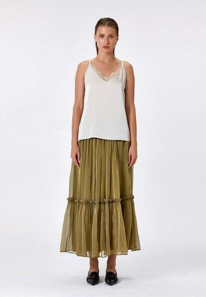 Femme debout, face à nous, portant un caraco blanc orné de dentelle, une jupe maxi plissée vert olive et des chaussures noires pointues.