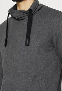 Sweat-shirt gris avec une capuche drapée, doté d'un cordon noir et d'une poche avant. Le matériau semble doux et texturé.