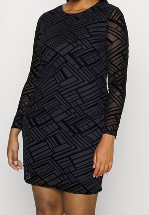 Kobieta ubrana w czarną sukienkę z długim rękawem, z przejrzystymi rękawami i geometrycznymi wzorami z aksamitu, stojąca na jednolitym jasnym tle.