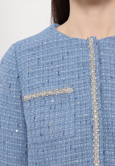 Hellblaue Tweedjacke mit strukturiertem Muster, dekorativem silbernen Besatz entlang der Frontöffnung und einer kleinen Tasche mit Verzierungen.
