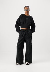 Svart cropped genser med løse ermer, sammen med vide svarte cargo-bukser. Modellen har på seg solbriller, og viser en moderne urban stil.
