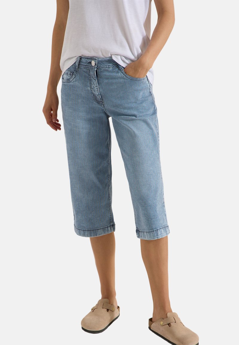 Personne portant un pantalon à rayures verticales bleu et blanc longueur genoux, un t-shirt blanc et des chaussures beiges à enfiler, debout avec une main dans la poche.