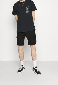 Černé grafické tričko s designem na hrudi, spárované s černými cargo šortkami. Na nohou má černobílé tenisky a bílé ponožky.