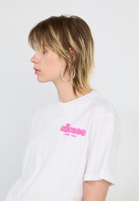 T-shirt blanc avec un logo "ellesse" rose sur la poitrine, à manches courtes et col rond. Le modèle affiche un profil latéral, portant de grandes boucles d'oreilles en argent.