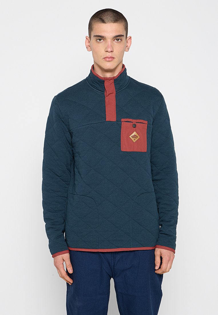 Pullover trapuntato in verde scuro con collo alto e dettagli color marsala. Presenta una tasca sul petto con dettaglio a bottone e tasche a marsupio.