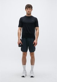 Svart atletisk t-shirt med kort ärm, grå shorts, vita strumpor och vita skor. Modell som håller en vattenflaska och står på en enfärgad bakgrund.
