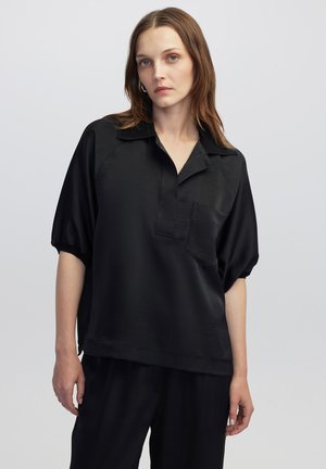 Camicetta nera a maniche corte con colletto, taschino anteriore e texture liscia. Indossata con pantaloni coordinati, caratterizzati da una vestibilità ampia.
