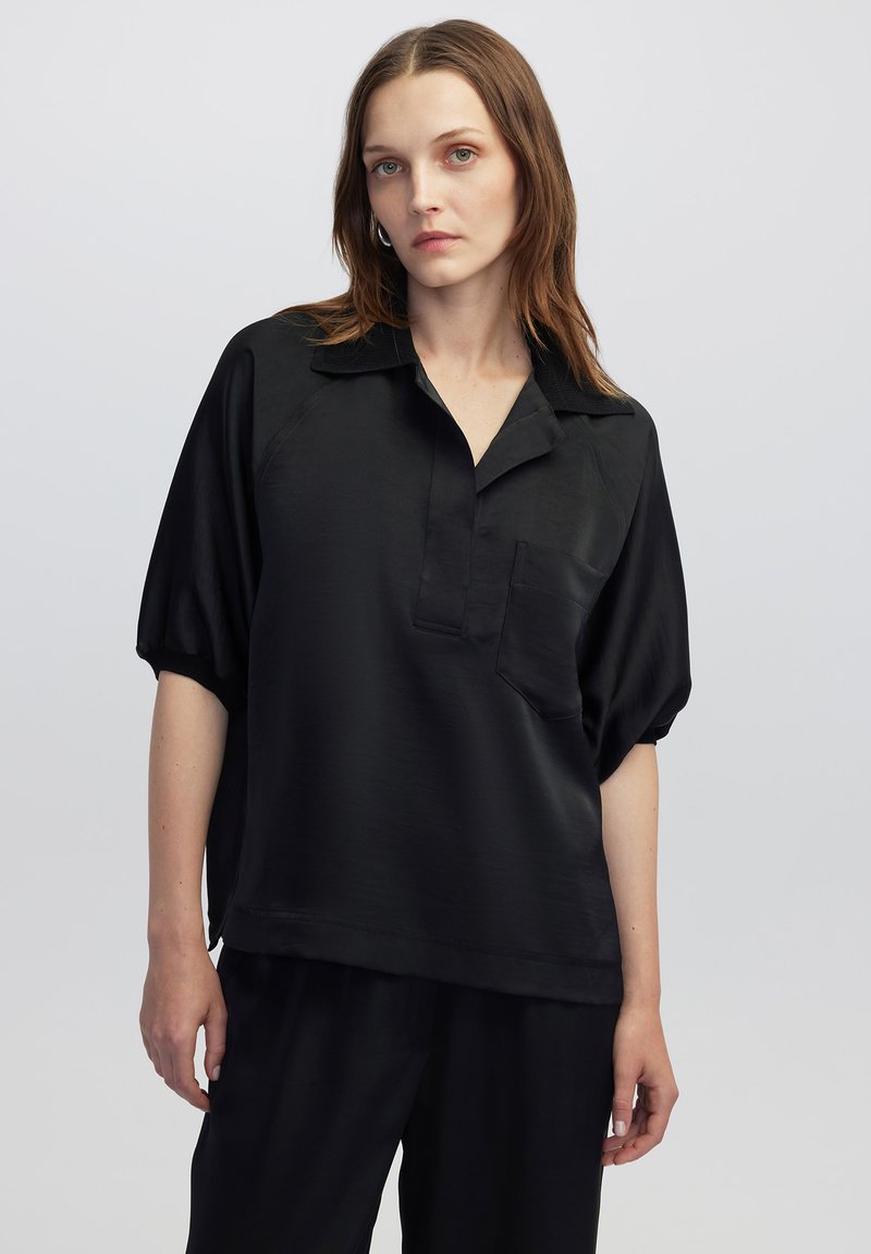 Camicetta nera a maniche corte con colletto, taschino anteriore e texture liscia. Indossata con pantaloni coordinati, caratterizzati da una vestibilità ampia.