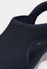 Κοντινό πλάνο μπλε σκούρου σανδάλι με υφή και πλεκτό σχέδιο και φαρδιά λουρί γύρω από τη φτέρνα και πλευρικό άνοιγμα για το πόδι.