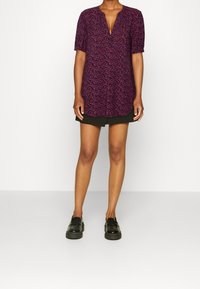 Vestido de manga corta, con un escote en V profundo en púrpura oscuro, con estampado floral en rojo y azul. Incluye un dobladillo curvado y se combina con zapatos negros.