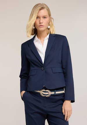 Blondynka ubrana w dopasowaną granatową marynarkę i spodnie, z białą koszulą i beżowym paskiem z podwójną klamrą, stoi na jasnym tle.