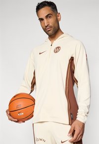 Crème hoodie met bruine zijaccenten, ritssluiting bij de nek en logo. De persoon houdt een oranje basketbal vast met zwarte details.