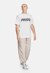 Biała bawełniana koszulka z czarnym napisem "PROSTO.", zestawiona z jasnobeżowymi spodniami typu cargo i białymi sportowymi trampkami z kolorowymi akcentami.