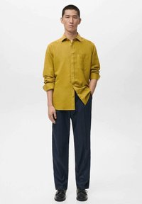Senfgelbes Hemd mit Knopfleiste und Brusttasche, kombiniert mit dunkelblauen Hosen. Das Outfit zeichnet sich durch eine lockere Passform und glatten Stoff aus.