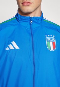 Blå sportjacka med vitt Adidas-logotyp och Italiens landslagssymbol, med zip-stängning och gröna och vita randiga detaljer.