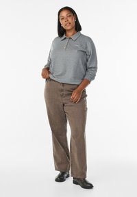 Femme aux cheveux noirs courts portant un polo gris à manches longues, un jean large marron et des bottines noires, se tenant devant un fond blanc.