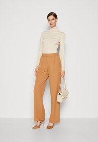 Beige eng anliegendes Rollkragenoberteil mit horizontalem Streifenmuster, kombiniert mit weit geschnittenen camel-farbenen Hosen und spitzen nude-farbenen Heels, während sie eine cremfarbene Handtasche hält.