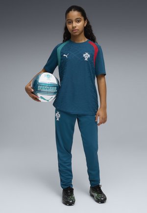 Ung kvindelig fodboldspiller i Portugals trøje, der holder en fodbold med Portugal og Puma logoer, stående mod en ens grå baggrund.