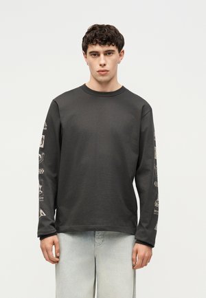 Νέος άνδρας με κοντά σγουρά μαλλιά που φοράει μαύρο μακρυμάνικο μπλουζάκι με γραφικά στα μανίκια και ανοιχτόχρωμα τζιν, στέκεται μπροστά σε απλό φόντο.