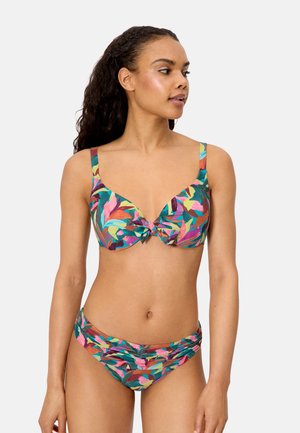 Mujer con cabello largo y rizado vistiendo un bikini con estampado tropical colorido, mirando hacia su izquierda sobre un fondo liso.