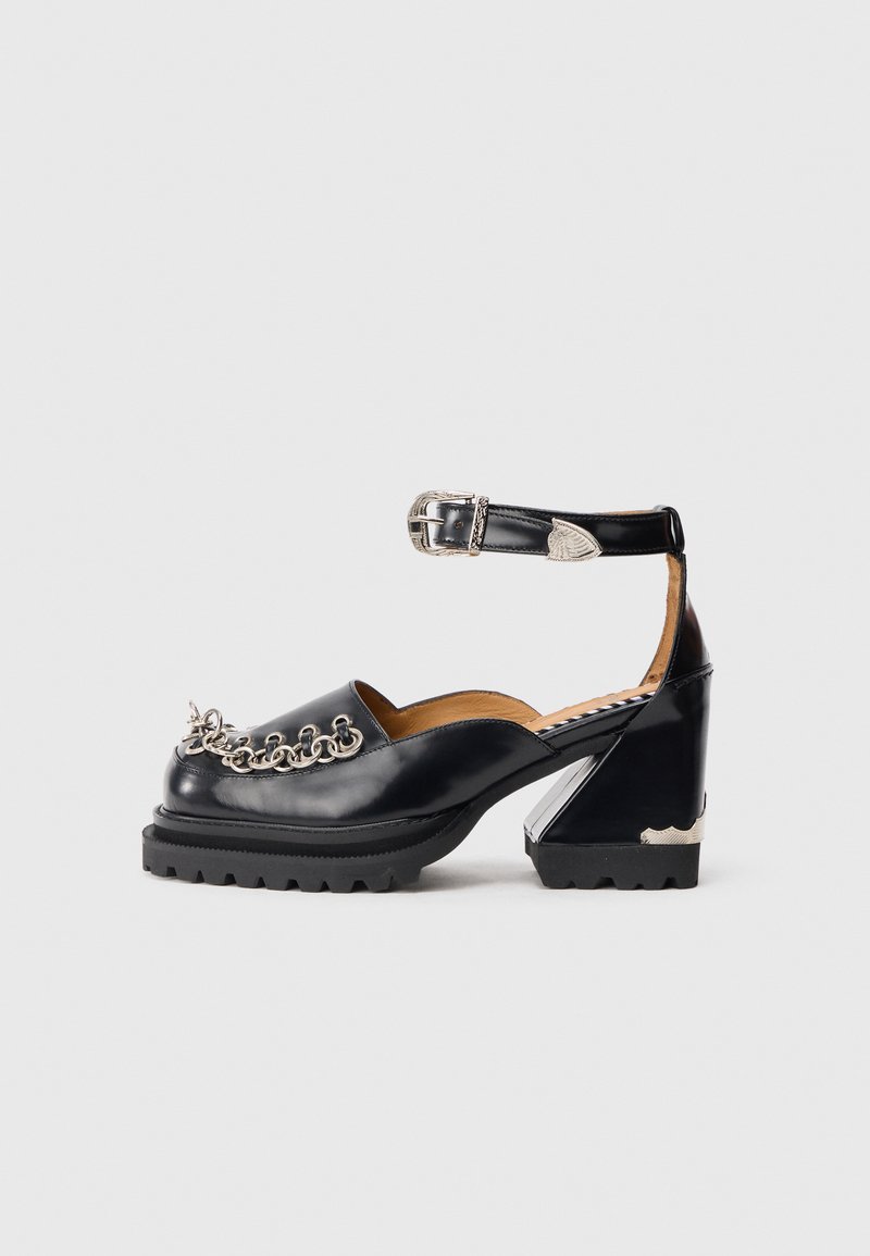 Black leather chunky-heeled shoe with silver chain detail on toe and ankle strap with silver buckle on white background.