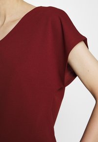 Gros plan d'une personne portant un haut à manches courtes rouge foncé avec un col en V sur un fond clair.