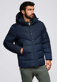 Giacca imbottita blu navy con cappuccio, dotata di chiusura con zip e motivi di cuciture diagonali, include tasche laterali e una texture morbida.