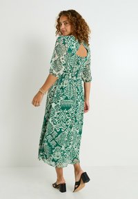 Robe verte à motifs avec une silhouette fluide, des manches mi-longues et un détail d'ouverture en forme de clé au dos. Portée avec des sandales à talons carrés noires.