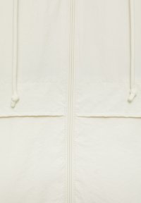 Biała lekka kurtka z frontowym zamkiem błyskawicznym, sznurkami do regulacji oraz teksturowaną tkaniną. Posiada dwie poziome kieszenie.