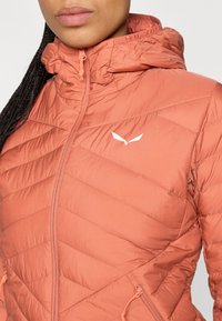 Casaco acolchoado pêssego com colarinho alto, fecho zip e dois bolsos com fecho zip. Apresenta material leve e resistente à água, e um logótipo.