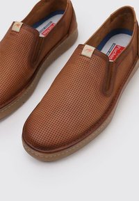 Zapatos marrones de cuero tipo mocasín con parte superior perforada, suela de goma y acentos beige. Presentan un pequeño distintivo con el logo en el lateral.