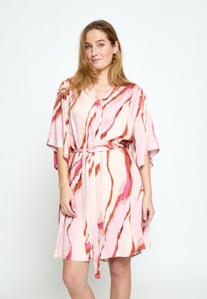 Frau trägt ein rosa kurzärmeliges Kleid mit roten und violetten abstrakten Streifen, die in der Taille gebunden sind, und steht vor einem schlichten weißen Hintergrund.