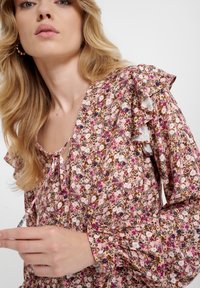 Kwiatowa bluzka w odcieniach różu i brązu, z falbanistymi rękawami, z wiązaniem przy szyi oraz mieszanym wzorem małych kwiatów i liści.