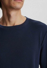Sötétkék, bordázott, hosszú ujjú póló kerek nyakkivágással, amely texturált felülettel és minimális varrásokkal rendelkezik a letisztult megjelenésért.