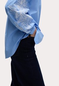 Blusa a righe blu chiaro con ricamo floreale bianco su una manica, abbinata a jeans di denim scuri. Presenta una vestibilità morbida e maniche arrotolate.
