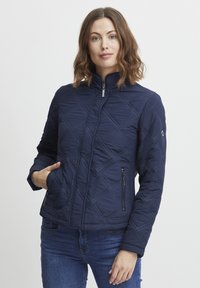 Jachetă matlasată de culoare albastru marin, cu guler în picioare, fermoar în față și buzunare laterale. Are cusături diagonale și un design ajustat.