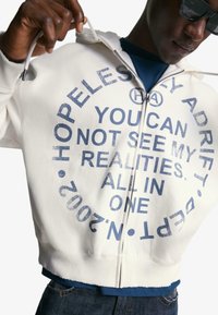 Weiße Zip-Hoodie mit blauer grafischer Schrift, die kreisförmig angeordnet ist. Dicke Bündchen und Saum mit gerippter Textur. Getragen über einem blauen Shirt.