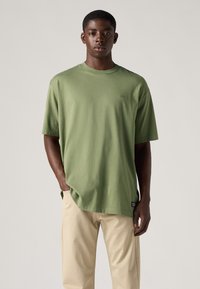 T-shirt in cotone verde oliva con maniche corte, vestibilità ampia, scollatura rotonda, con logo discreto sul petto, abbinato a pantaloni beige.