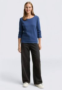 Blauw langärmelig topje met een ronde hals en geribbelde structuur, gecombineerd met zwarte broek met wijd uitlopende pijpen en kleine witte stippen, afgewerkt met witte sneakers.